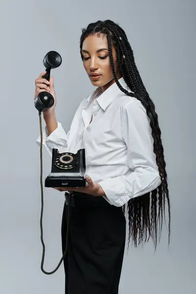 A young woman with long black braids poses holding a vintage rotary phone against a gray backdrop. — Stock Photo