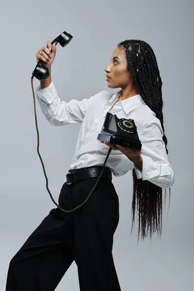A young African American woman with braids, wearing a white shirt and black pants, holds a vintage rotary phone while looking up. — Stock Photo