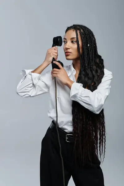 Une jeune femme aux longues tresses pose en chemise blanche sur fond gris, tenant un téléphone vintage à l'oreille. — Stock Photo