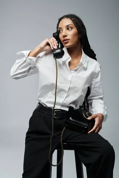 A young African American woman in a white shirt and black pants sits and talks on a vintage rotary phone. — Stock Photo