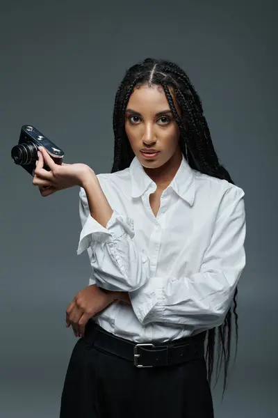 A young woman with long braids wears a white shirt and poses with a camera in hand against a grey backdrop. — Stock Photo