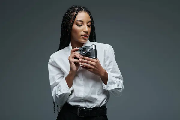 A young woman in a white shirt stands against a grey background, holding a camera in her hands, focused on the lens. — Stock Photo