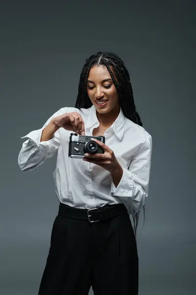 A young African American woman in a white shirt and black pants poses against a gray background, holding a vintage camera and smiling. — Stock Photo