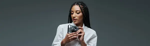 A young woman with braided hair poses against a grey background, holding a camera in her hands. — Stock Photo