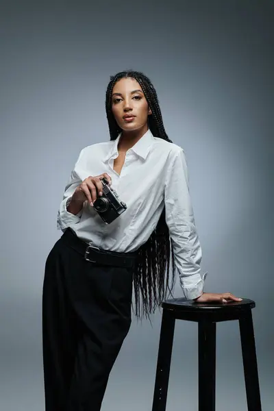 Une femme élégante avec de longues tresses s'appuie sur un tabouret tout en tenant une caméra. — Photo de stock