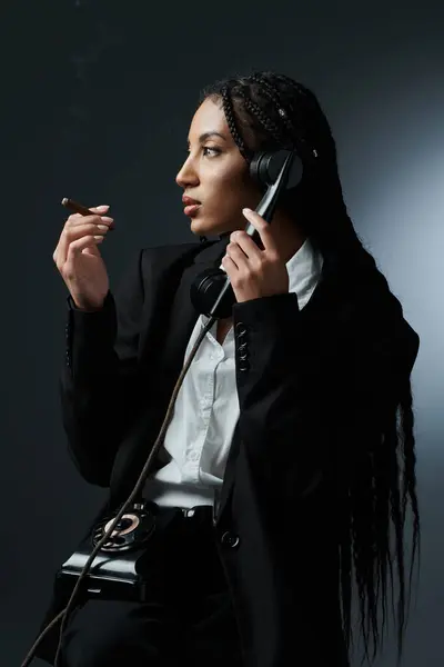 A stylish young woman in a white shirt and black blazer sits with a vintage phone and a lit cigar, looking thoughtfully into the distance. — Stock Photo