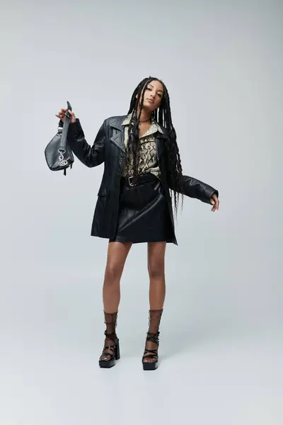 A young woman in a black leather jacket and skirt poses confidently with handbag against a grey backdrop, fashion and style — Stock Photo