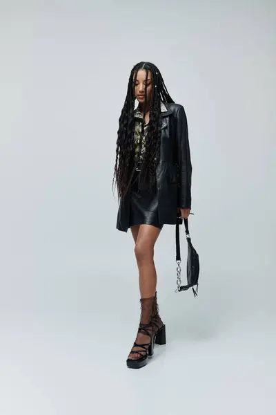 A young woman in a black leather jacket and skirt poses confidently against a grey background, her long braids cascading down her back. — Stock Photo