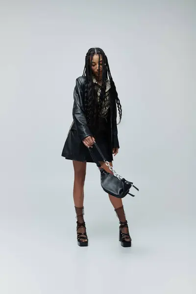 A young woman in a black leather jacket, skirt, and shoes poses with a black handbag. Her long black hair cascades down her back. — Stock Photo