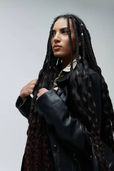 A young woman with long braids and a stylish black leather jacket poses against a grey backdrop, radiating confidence and individuality. — Stock Photo