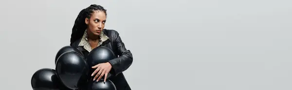 In her 20s, a chic Black woman in black leather poses with black balloons against a grey backdrop, her gaze intense and intriguing — Stock Photo
