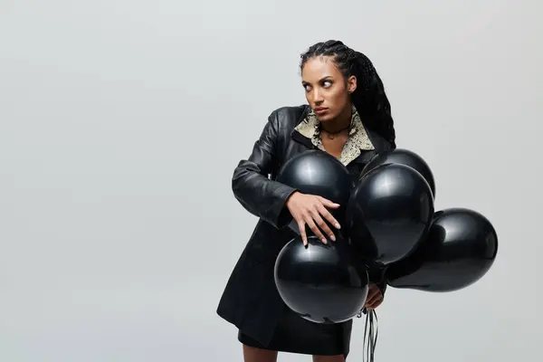 A stylish African American woman in a black leather outfit holds a bunch of black balloons while looking away from the camera. — Stock Photo