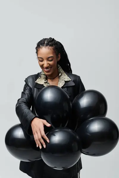 Une jeune femme vêtue d'une veste en cuir noir et d'une chemise à imprimé animal pose avec un tas de ballons noirs sur un fond gris. — Stock Photo