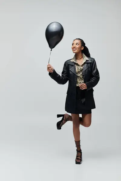 A young woman in a black leather jacket and skirt poses with a black balloon, looking up dreamily. — Stock Photo