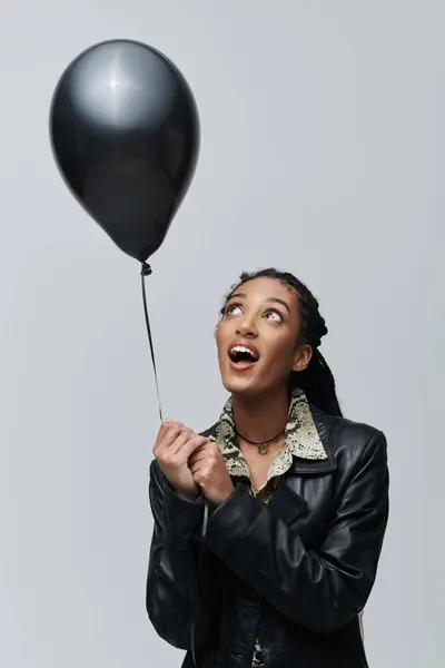 A young woman dressed in a black leather jacket and animal print shirt looks up at a black balloon in her hand. — Stock Photo