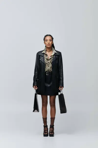 Une jeune femme élégante pose dans une veste et une jupe en cuir noir, avec une chemise à motifs, tenant des sacs à provisions. — Photo de stock