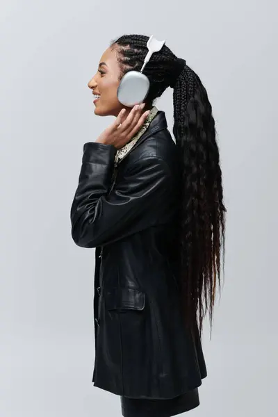 Una giovane donna in giacca di pelle nera e camicia stampa animalier posa su uno sfondo grigio, cuffie accese, ascoltando musica. — Foto stock