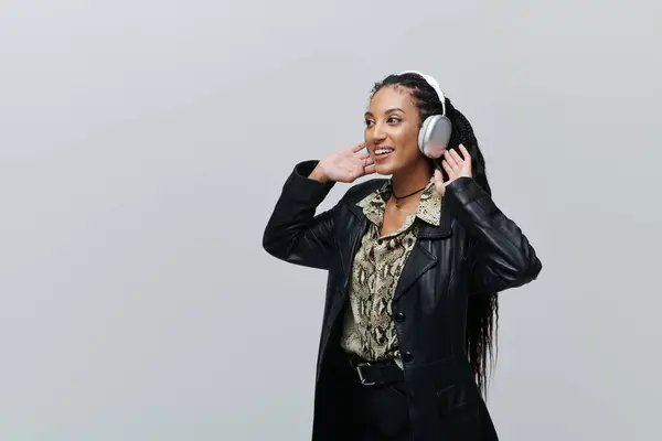 Una giovane donna afroamericana in giacca di pelle nera e camicia stampa animalier sorride ascoltando musica con le cuffie. — Foto stock