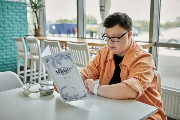A man with Down syndrome sits at a table in a cafe, carefully reviewing the menu and choosing what to eat. — Stock Photo