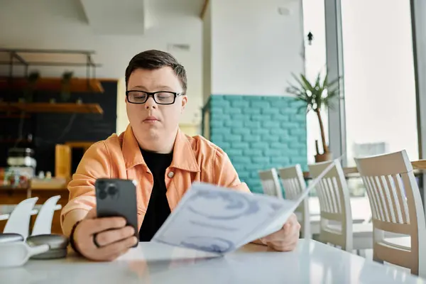 Ein Mann mit Down-Syndrom sitzt an einem Cafétisch, betrachtet eine Speisekarte und benutzt sein Handy. — Stockfoto