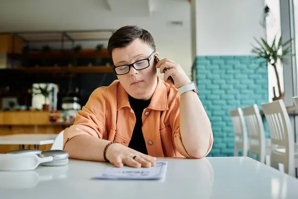 A man with down syndrome is sitting at a cafe table looking at a menu while talking on his phone. — Stock Photo