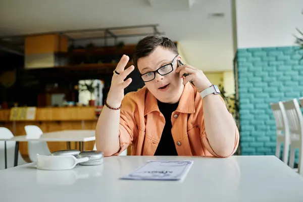 A man with Down Syndrome, wearing glasses and a peach jacket, sits at a cafe table, speaking on his phone and looking at the menu. — Stock Photo