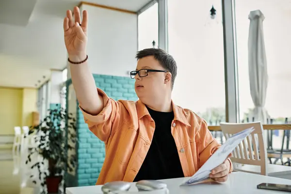 Un homme trisomique s'assoit à une table dans un café, tenant un menu et levant la main pour commander. — Photo de stock