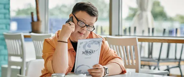 A man with Down syndrome sits at a cafe table, thoughtfully reviewing the menu and speaking on the phone. — Stock Photo