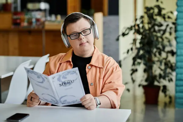 Ein Mann mit Down-Syndrom sitzt an einem Cafétisch und blickt gelassen über die Speisekarte. Brille und Kopfhörer tragen, Unabhängigkeit genießen — Stockfoto