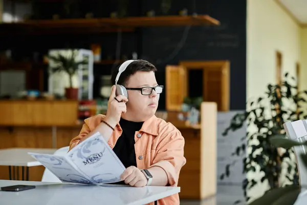 A man with Down syndrome, wearing headphones, sits at a cafe table and carefully considers the menu options. — Stock Photo