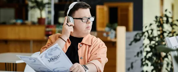 A man with Down syndrome wearing glasses and headphones reads the menu while sitting at a table in a cafe. — Stock Photo