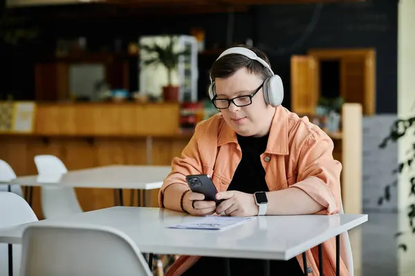 A young man with Down syndrome sits at a cafe table, headphones on, looking at his phone. — Stock Photo