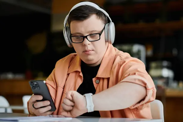 A young man with Down syndrome works remotely while wearing headphones and checking his smartwatch. — Stock Photo