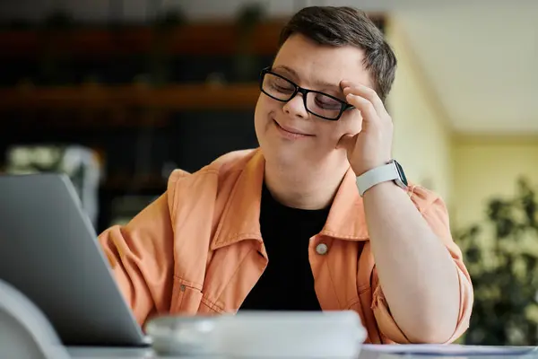 A man with Down syndrome works on his laptop while sitting in a cafe. He is wearing glasses and a peach-colored jacket. — Stock Photo