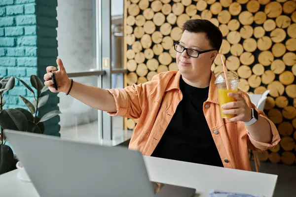 A man with Down syndrome sits at a cafe table, working on a laptop while holding a glass of orange juice. — Stock Photo