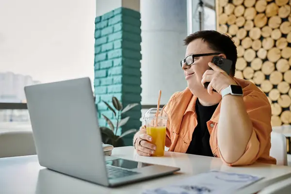 A man with Down syndrome enjoys a bright orange juice while working remotely on his laptop in a cafe. — Stock Photo