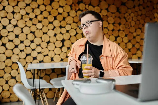A man with Down syndrome, wearing glasses and a peach-colored jacket, works on a laptop at a cafe, holding a glass of orange juice. — Stock Photo
