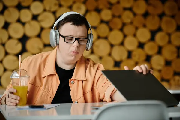 A man with Down syndrome, wearing headphones, works remotely on a laptop at a cafe while enjoying a drink. — Stock Photo