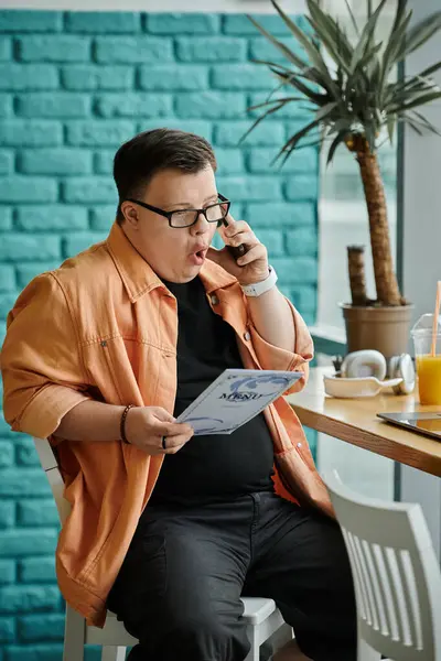 A man with Down Syndrome sits in a cafe, talking on his phone while looking at a document. — Stock Photo