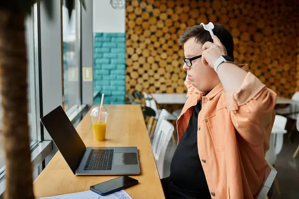 A man with Down syndrome adjusts his headphones while working remotely on his laptop at a cafe. — Stock Photo