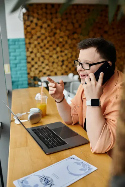 A man with Down syndrome works remotely on his laptop at a cafe, talking on his phone. — Stock Photo