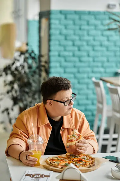 A man with Down syndrome sits at a cafe table, wearing glasses and a peach jacket, enjoying a slice of pizza and a glass of juice. — Stock Photo