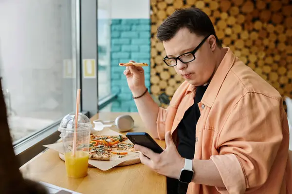 A man with Down syndrome sits at a cafe table, enjoying a slice of pizza and using his phone. — Stock Photo