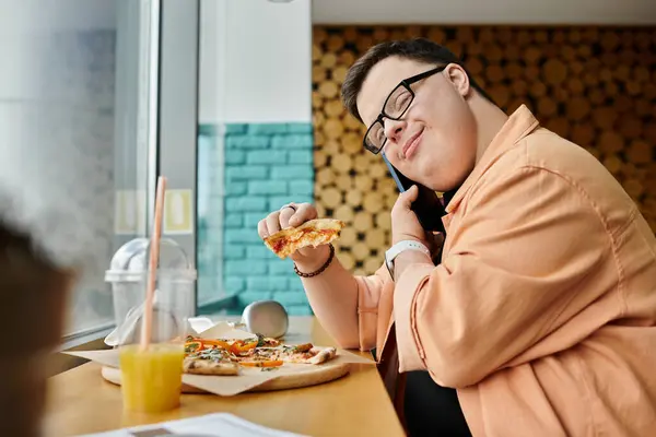 A man with Down syndrome enjoys a slice of pizza at a cafe, talking on his phone while savoring the delicious meal. — Stock Photo