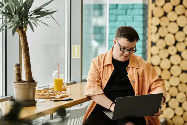 A man with Down syndrome sits in a cafe, enjoying a tasty pizza and working on his laptop. — Stock Photo
