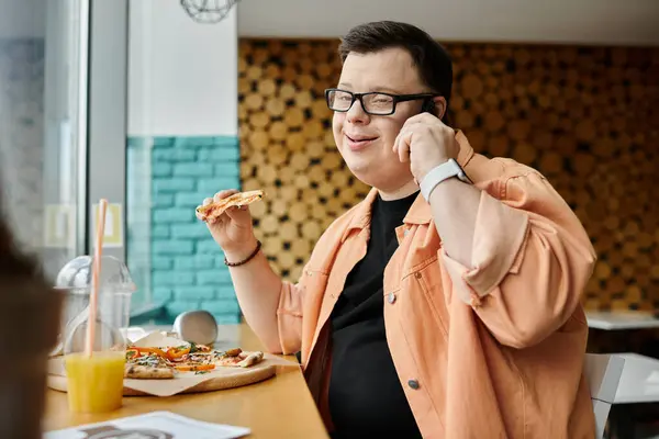 A man with Down syndrome enjoys a slice of pizza at a cafe, while taking a phone call. — Stock Photo