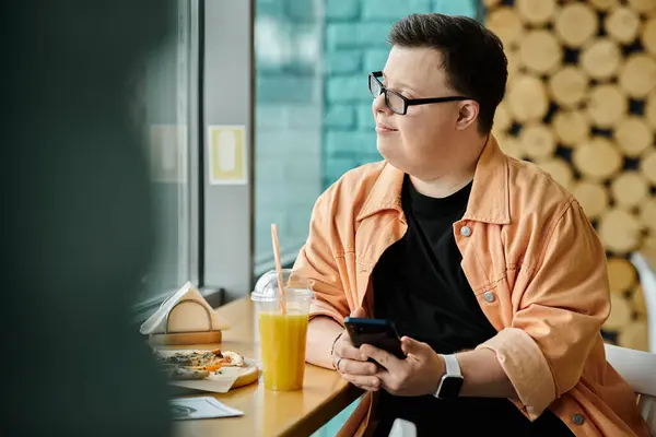 A man with Down syndrome, wearing glasses and an orange jacket, smiles as he enjoys a slice of pizza while sitting at a cafe table. — Stock Photo