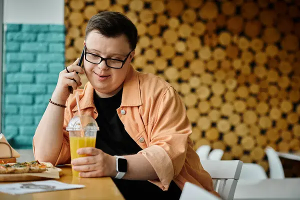 Un homme trisomique s'assoit dans un café, bavardant joyeusement sur son téléphone tout en dégustant une part de pizza et une boisson rafraîchissante.. — Photo de stock