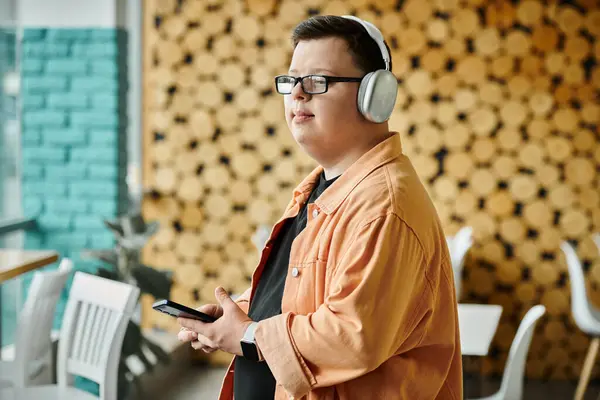A man with Down syndrome wearing headphones and glasses stands in a cafe, holding his phone. — Stock Photo