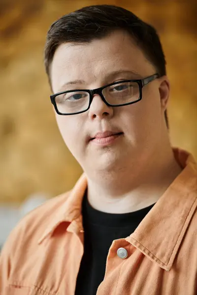 A young man with Down syndrome wears glasses and a peach-colored shirt as he looks into the camera. — Stock Photo